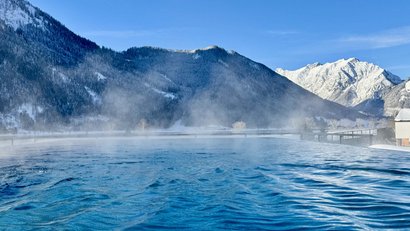 Heated outdoor pool with steam in front of snow-covered mountains under clear sky
