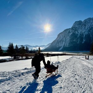 Active family holidays at Lake Achensee Person pulling another on a sled through a snowy landscape on a sunny day