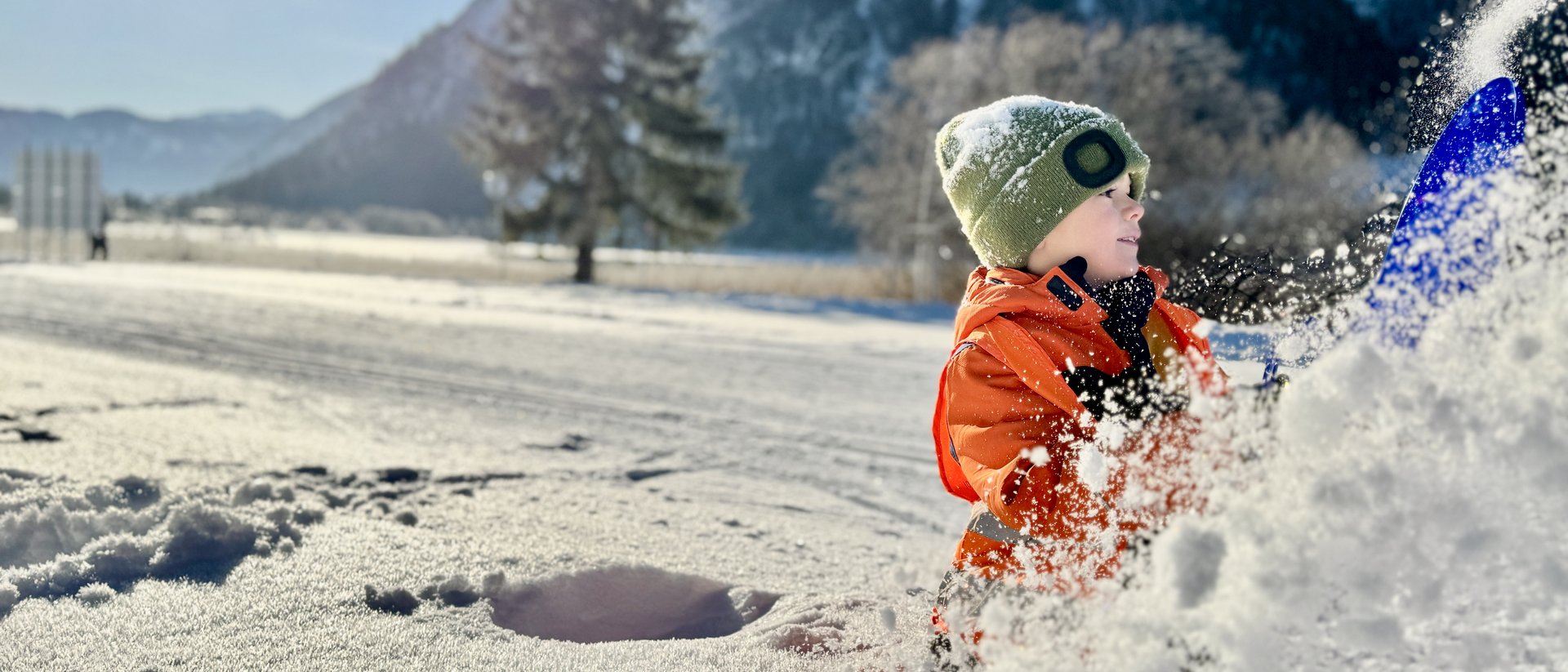 Child playing in snow with blue shovel in front of snowy mountains
